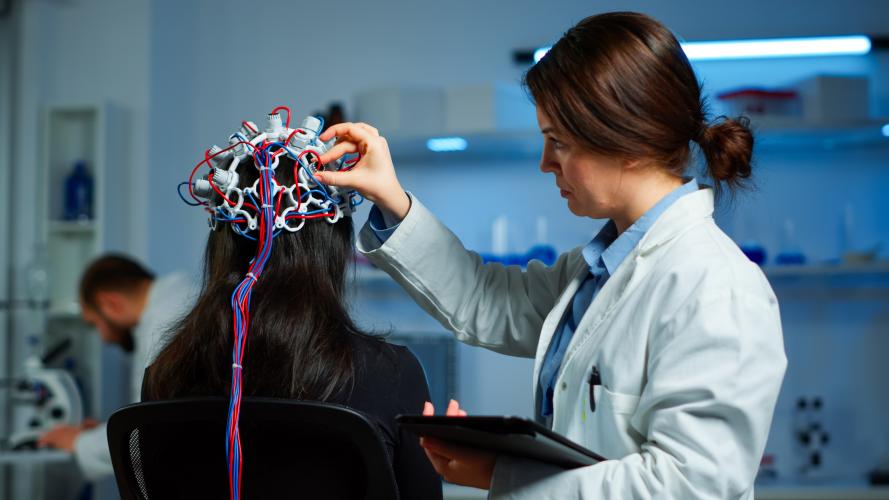Researcher fits and adjusts an EEG brain-monitoring cap with electrode leads on a seated participant in a neuroscience laboratory while holding a tablet.